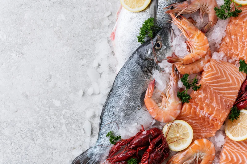 Assorted fresh raw seafood arranged on ice with lemon and parsley along the right side of a white textured surface