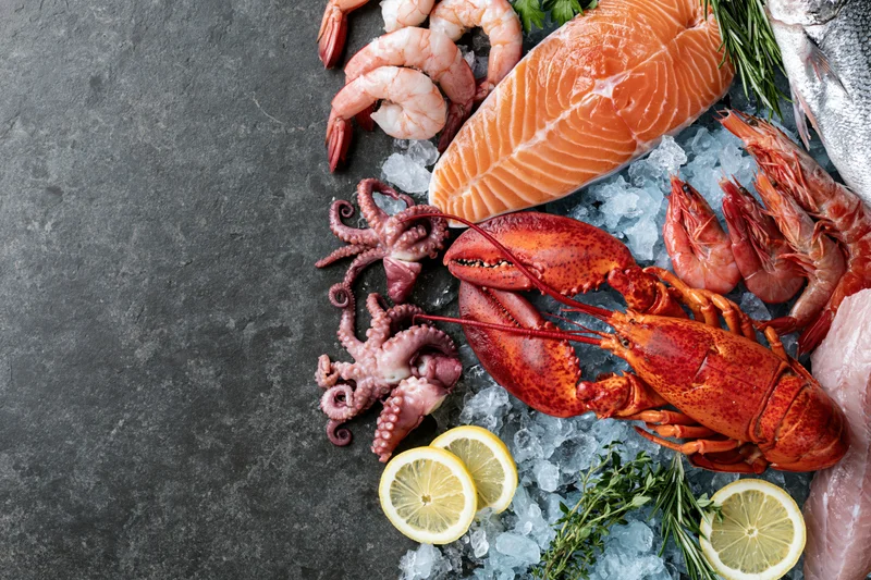 Assorted fresh seafood including salmon, lobster, shrimp and octopus arranged on crushed ice with lemon and herbs on dark surface