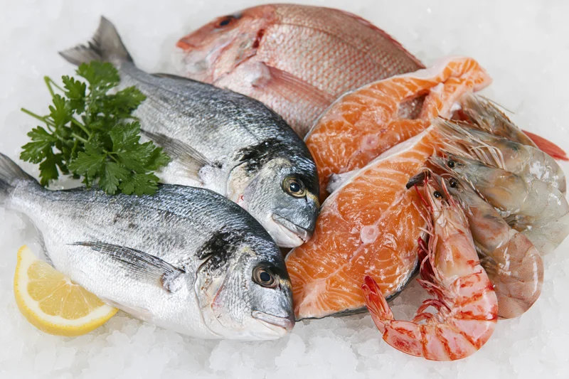 Assorted fresh seafood including whole fish, salmon fillets, and shrimp arranged on crushed ice with lemon and parsley