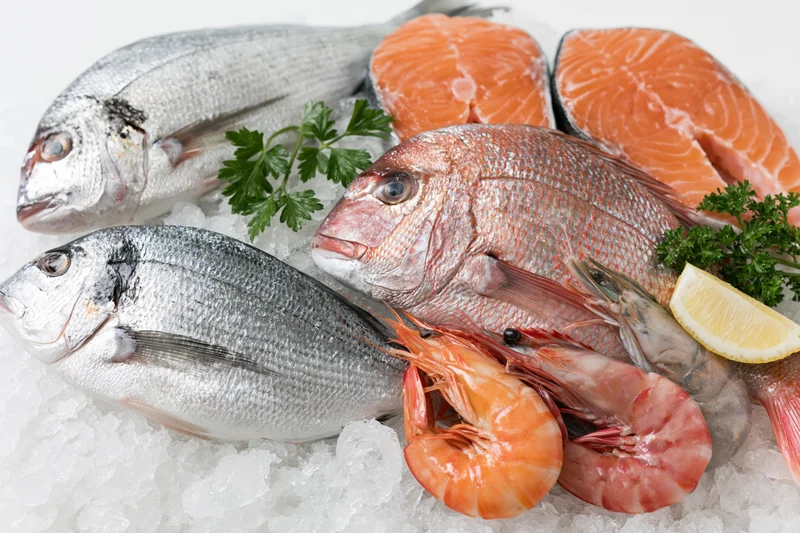 Assorted fresh seafood including whole fish, salmon fillets, and shrimp arranged on crushed ice with parsley and lemon slices