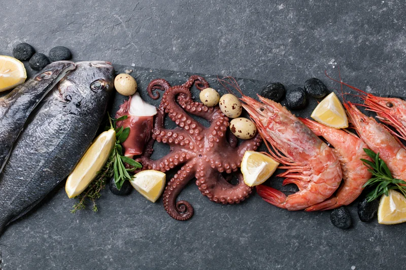 Raw seafood including octopus, shrimp, and fish arranged with lemon, herbs, and quail eggs on a dark slate surface