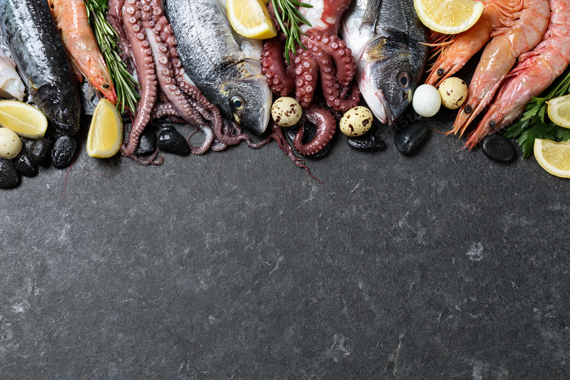 Raw seafood including fish, octopus and shrimp arranged along the top edge of a dark stone surface with lemon, herbs and quail eggs, leaving copyspace below