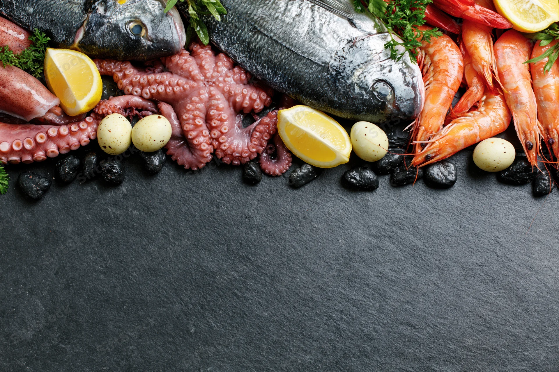 Assorted raw seafood arranged along the top edge of a dark slate surface with lemon, garlic, and herbs