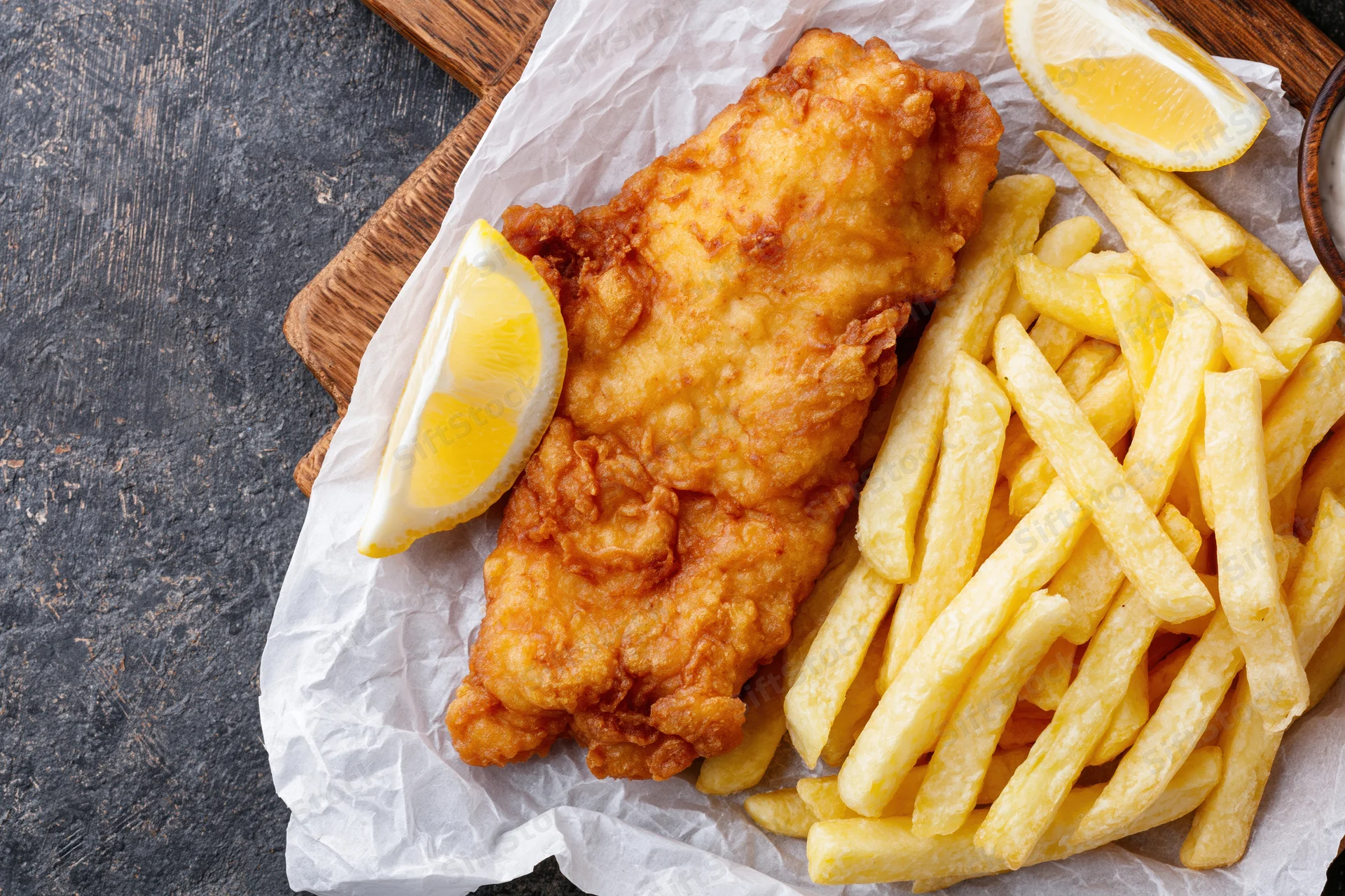 Battered fish fillet with golden french fries and lemon wedges on parchment paper over wooden board