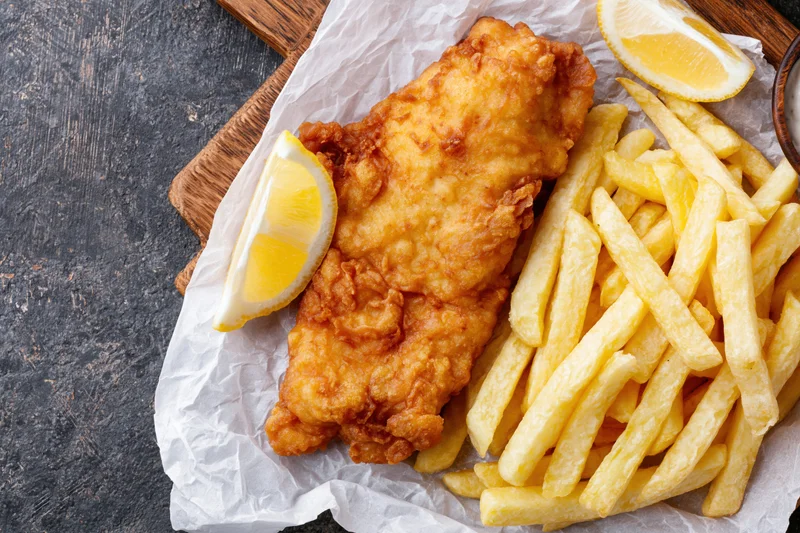 Battered fish fillet with golden french fries and lemon wedges on parchment paper over wooden board