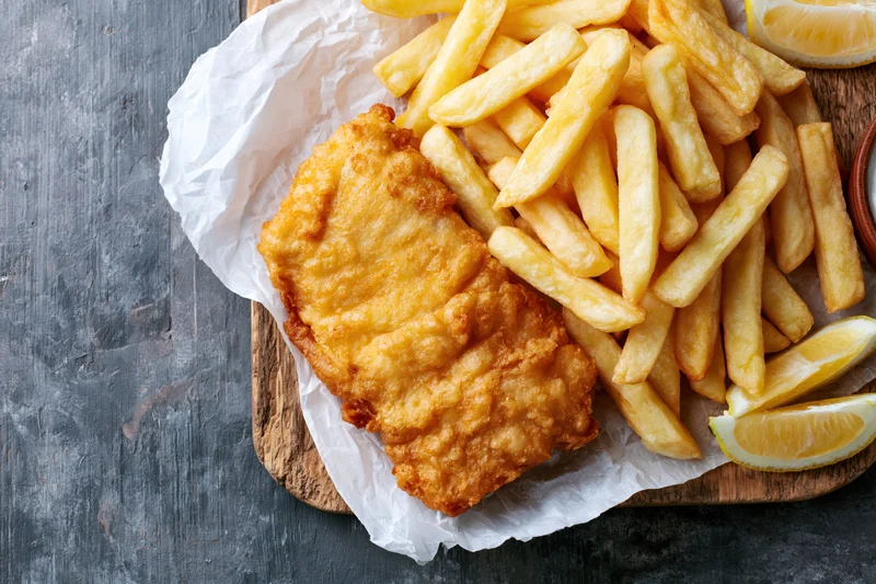Battered fish fillet with golden french fries and lemon wedges on a wooden board lined with paper