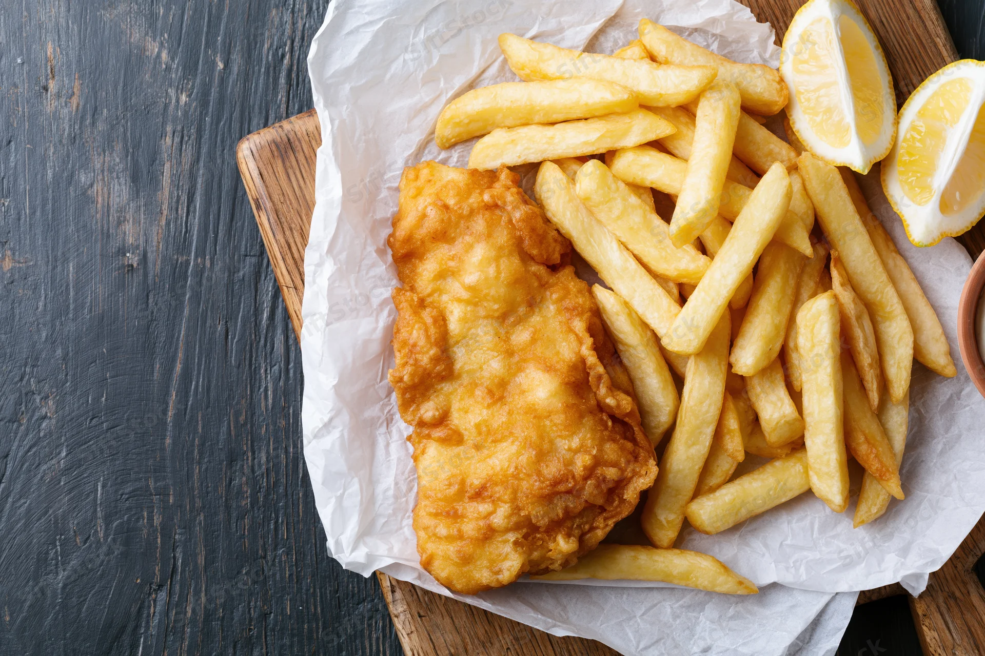Battered fish fillet with golden french fries and lemon wedges on paper-lined wooden board