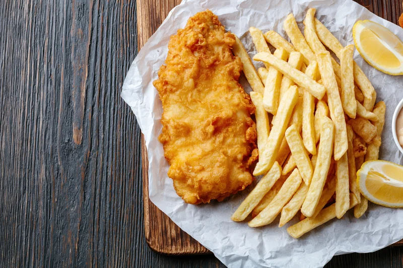 Battered fish fillet and golden french fries served on parchment paper on a wooden board