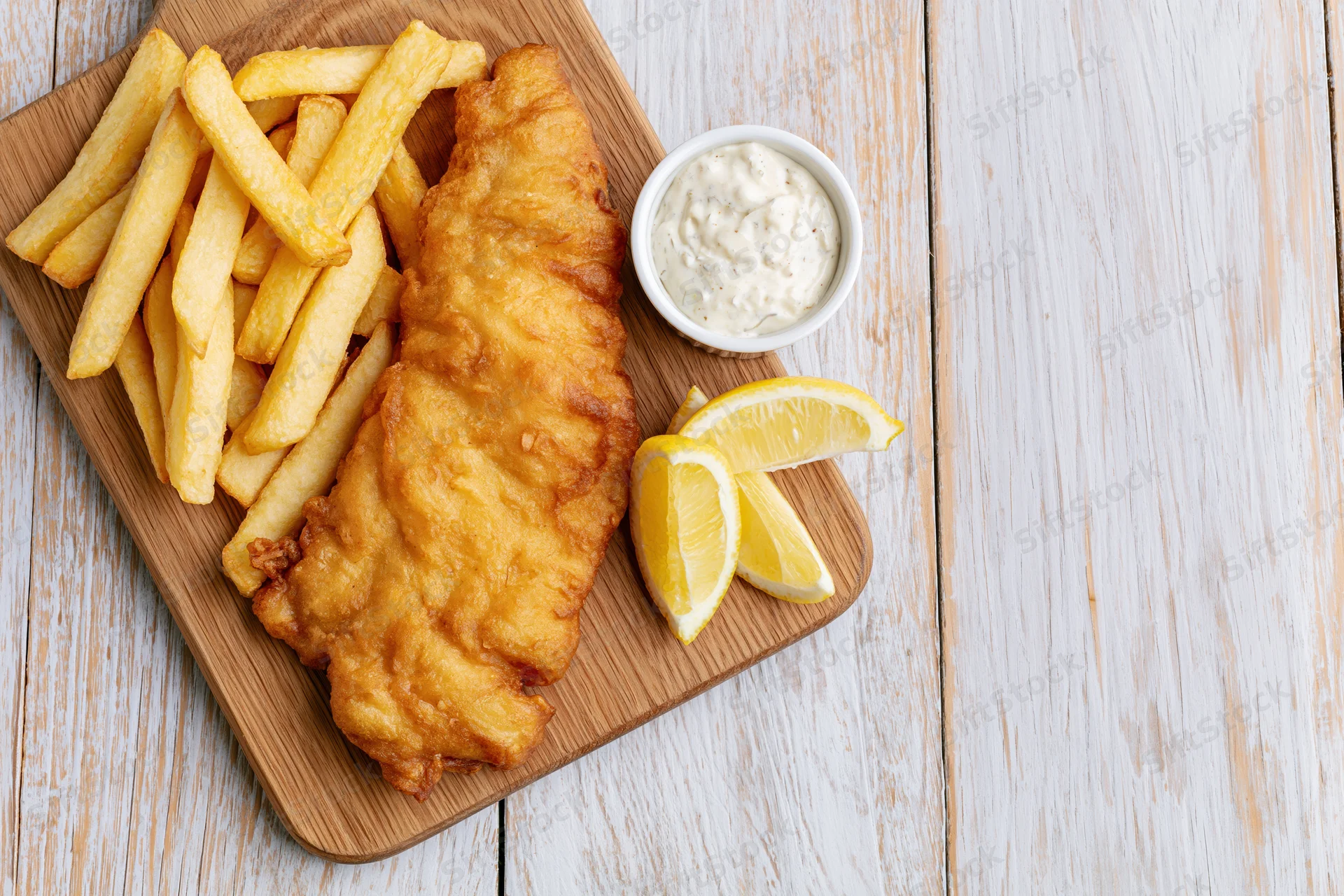 Beer-battered fish fillet and golden french fries served on a wooden board with lemon wedges and tartar sauce on a white wooden table