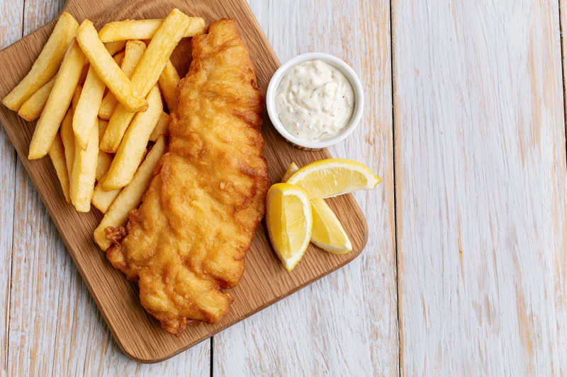 Beer-battered fish fillet and golden french fries served on a wooden board with lemon wedges and tartar sauce on a white wooden table