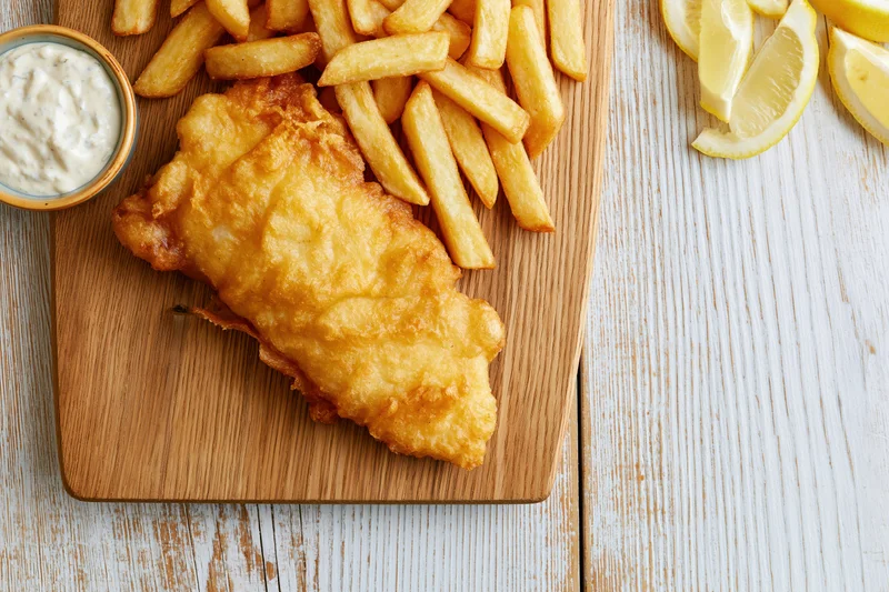 Beer-battered fish fillet with golden french fries on a wooden board beside lemon wedges and tartar sauce