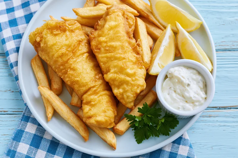 Fish and fries served on a white plate with lemon wedges and tartar sauce on a blue wooden table with a checkered napkin