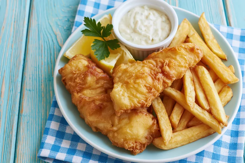 Fried fish fillets and french fries on a plate with tartar sauce and lemon wedges on a blue checkered napkin over a wooden table
