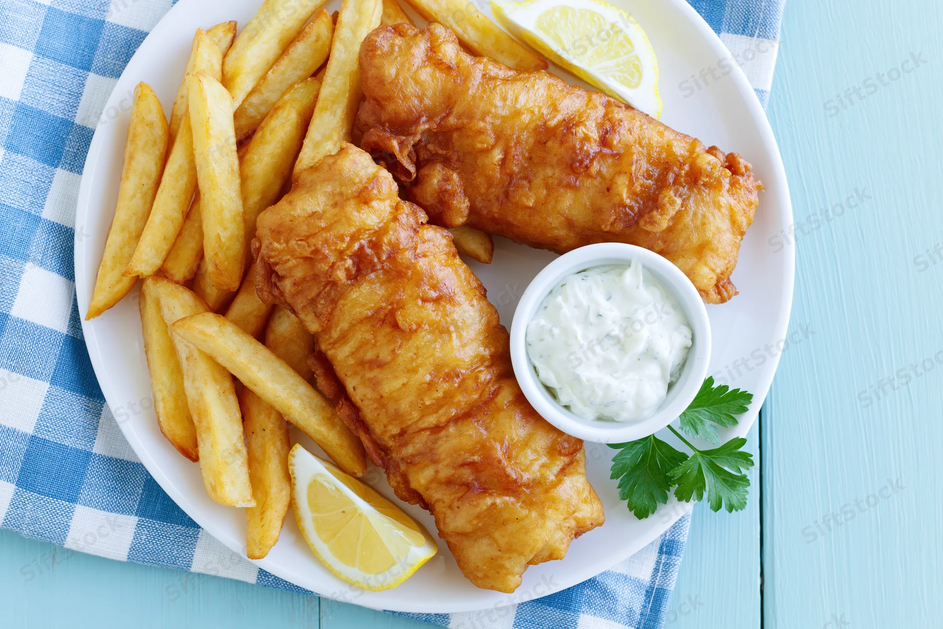 Two beer-battered fish fillets with french fries and tartar sauce on a white plate over a blue checkered napkin