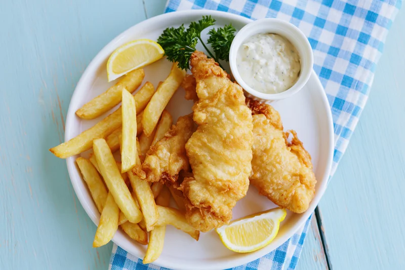 Beer-battered fish fillets and golden french fries served on a white plate with lemon wedges and tartar sauce on a blue checkered napkin over a wooden table