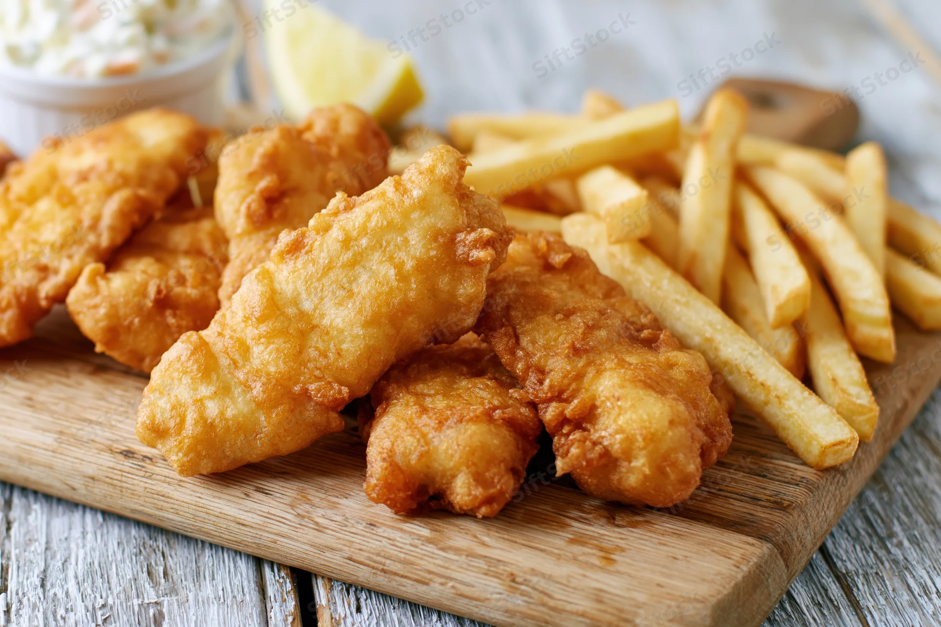Fried fish pieces and golden french fries served on a wooden cutting board with lemon and coleslaw