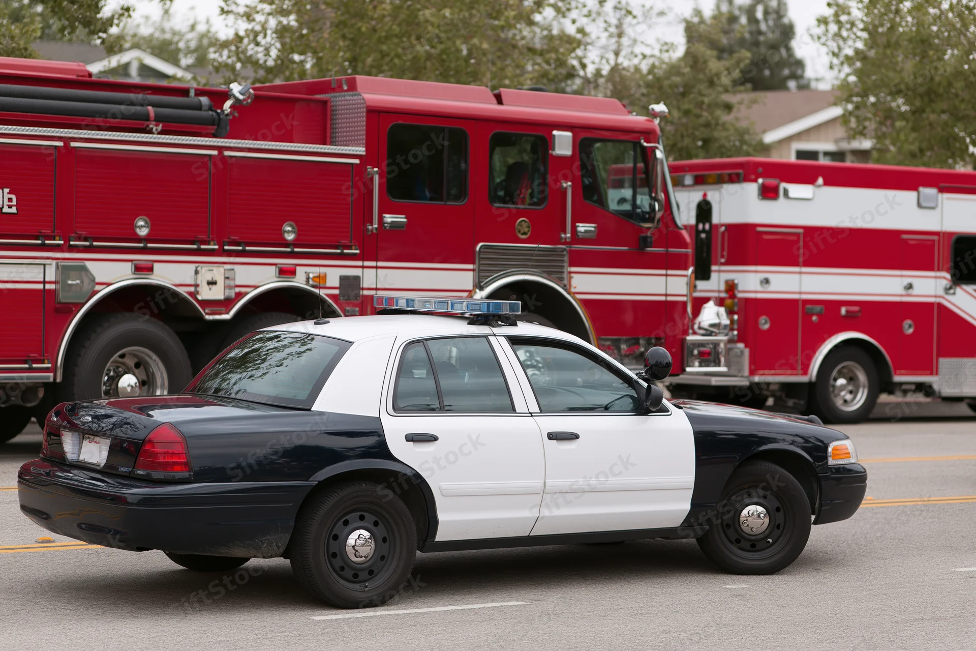 Black and white police cruiser parked in front of red fire trucks on asphalt street