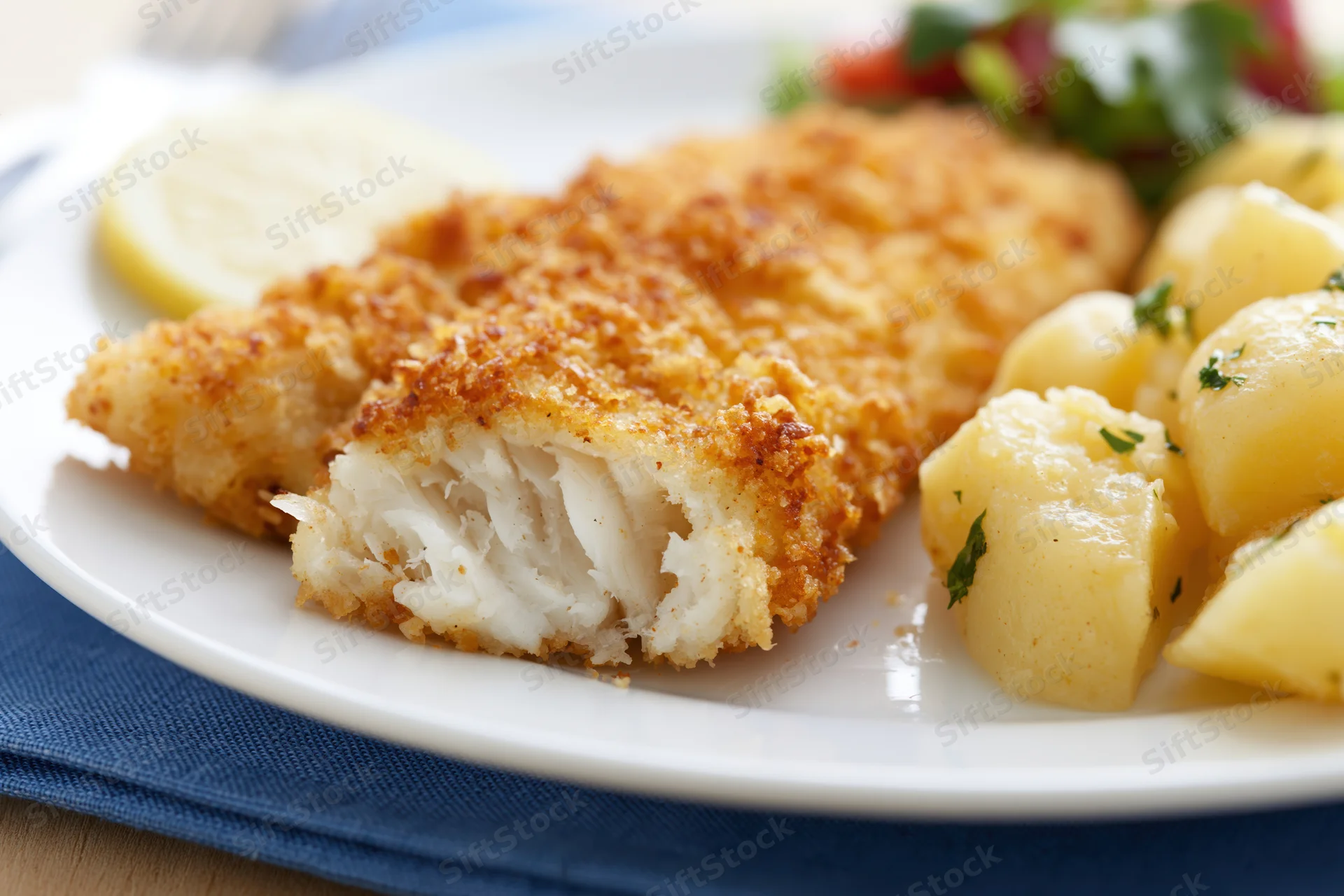 Breaded fried fish fillet with flaky white interior served on a plate with boiled potatoes and lemon garnish