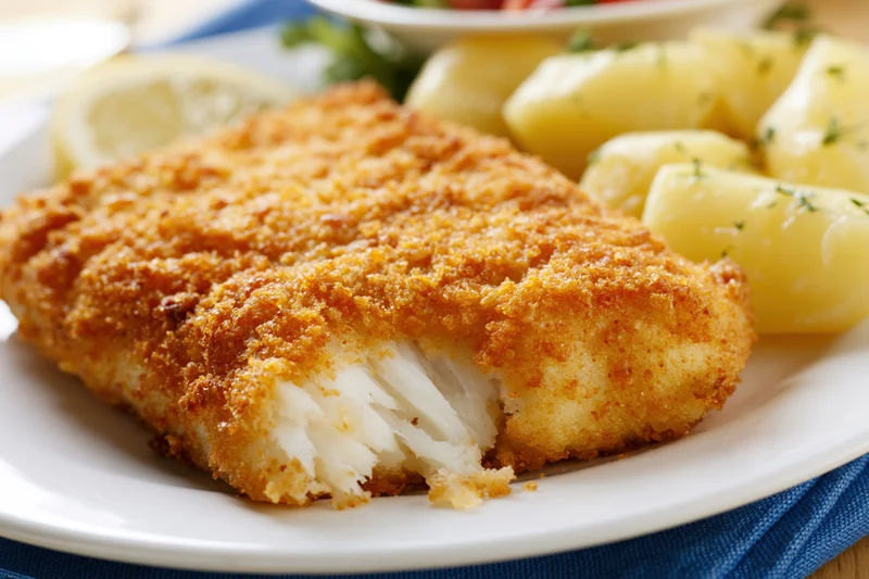 Breaded fried fish fillet with flaky white interior on a plate with potatoes and lemon garnish