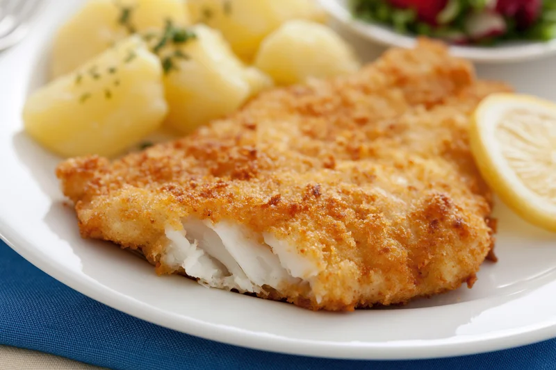 Breaded fried fish fillet with flaky white interior on a white plate with boiled potatoes and lemon slice