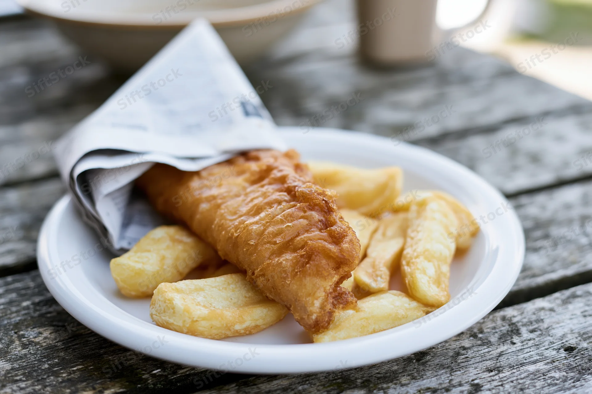Golden fried fish and chips on a white ceramic plate wrapped in newspaper on a wooden table