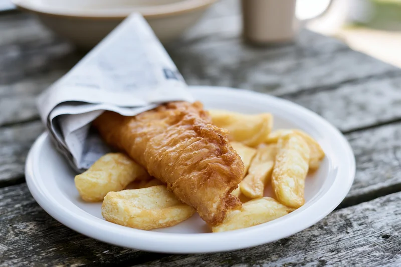 Golden fried fish and chips on a white ceramic plate wrapped in newspaper on a wooden table