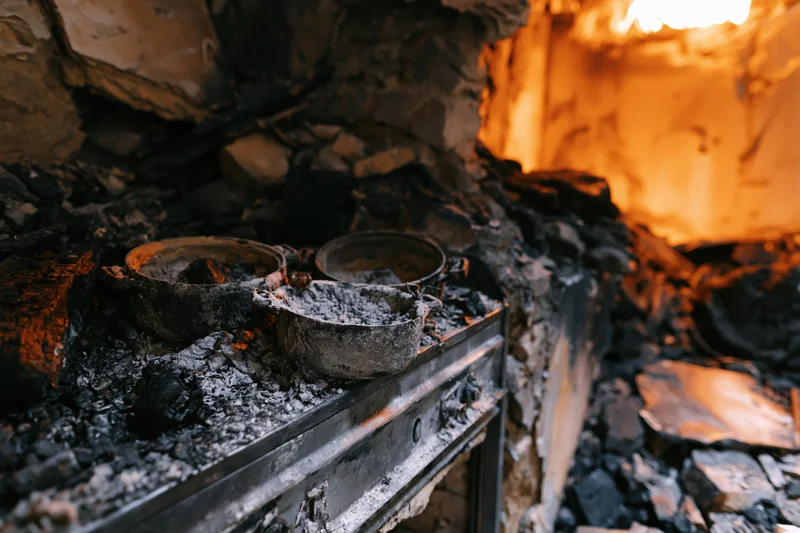 Burned kitchen interior with charred debris and ash-covered stove and pots after fire damage