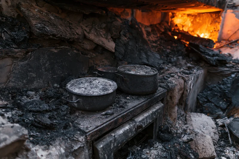 Burned kitchen interior with charred debris and cooking pots on a ruined table near a fire-damaged wall