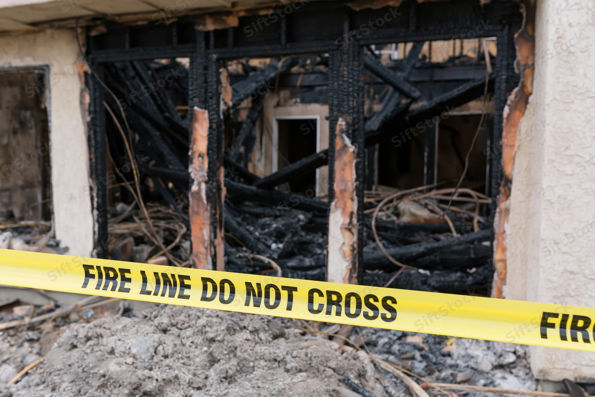 Burned residential building facade with charred remains and yellow fire line tape across rubble