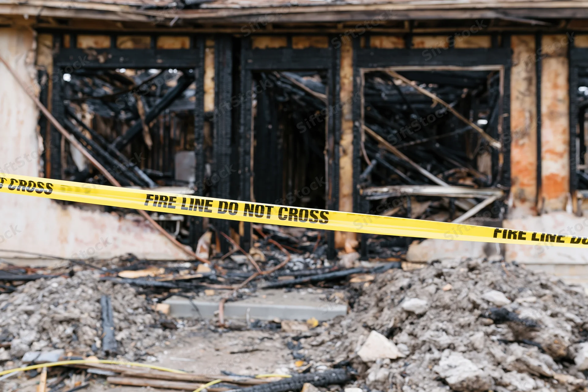 Burned residential building facade with charred wood frame and rubble, cordoned by yellow fire line tape