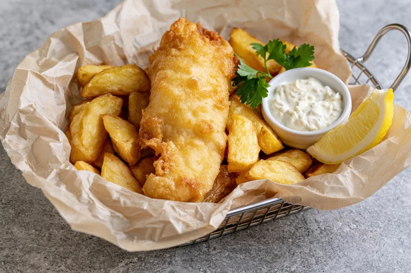 Fried fish and chips served in a metal basket lined with paper on a gray surface