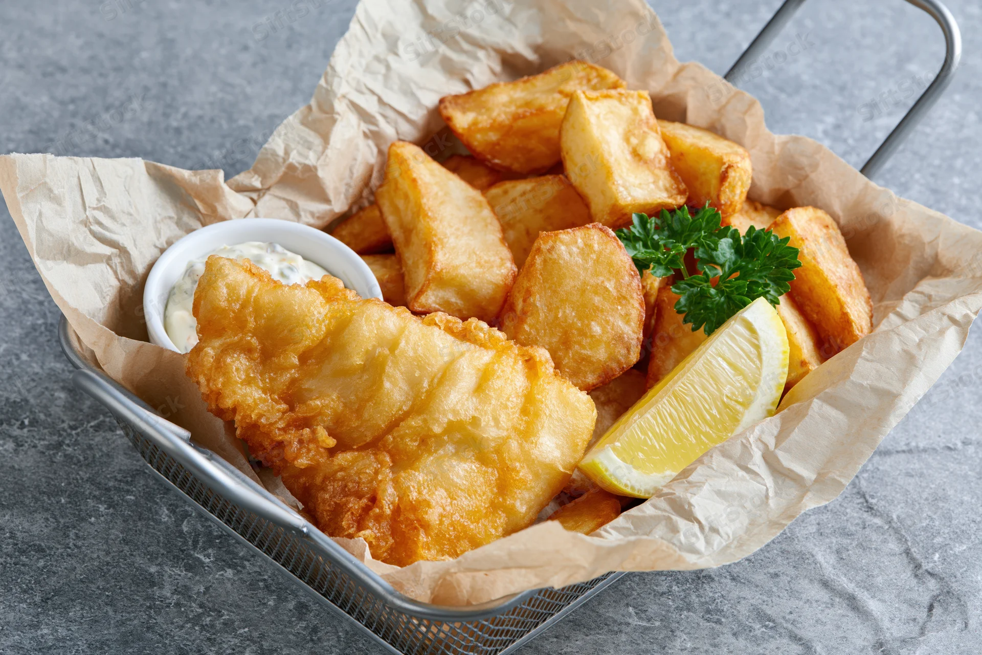 Fried fish and chips in a metal basket lined with paper, garnished with lemon and parsley, served with tartar sauce