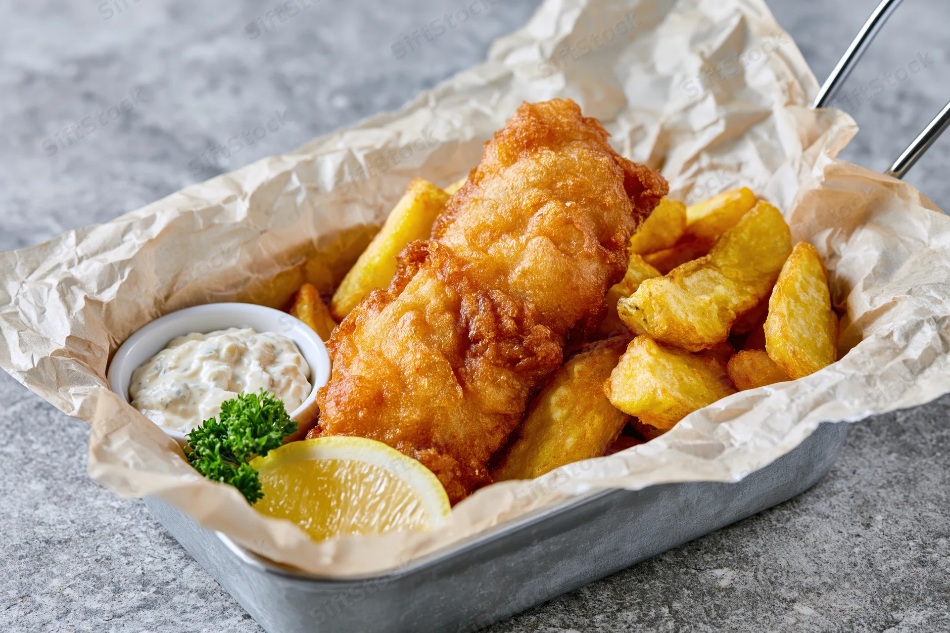Fried fish and chips in a metal basket lined with paper, served with tartar sauce and lemon slice