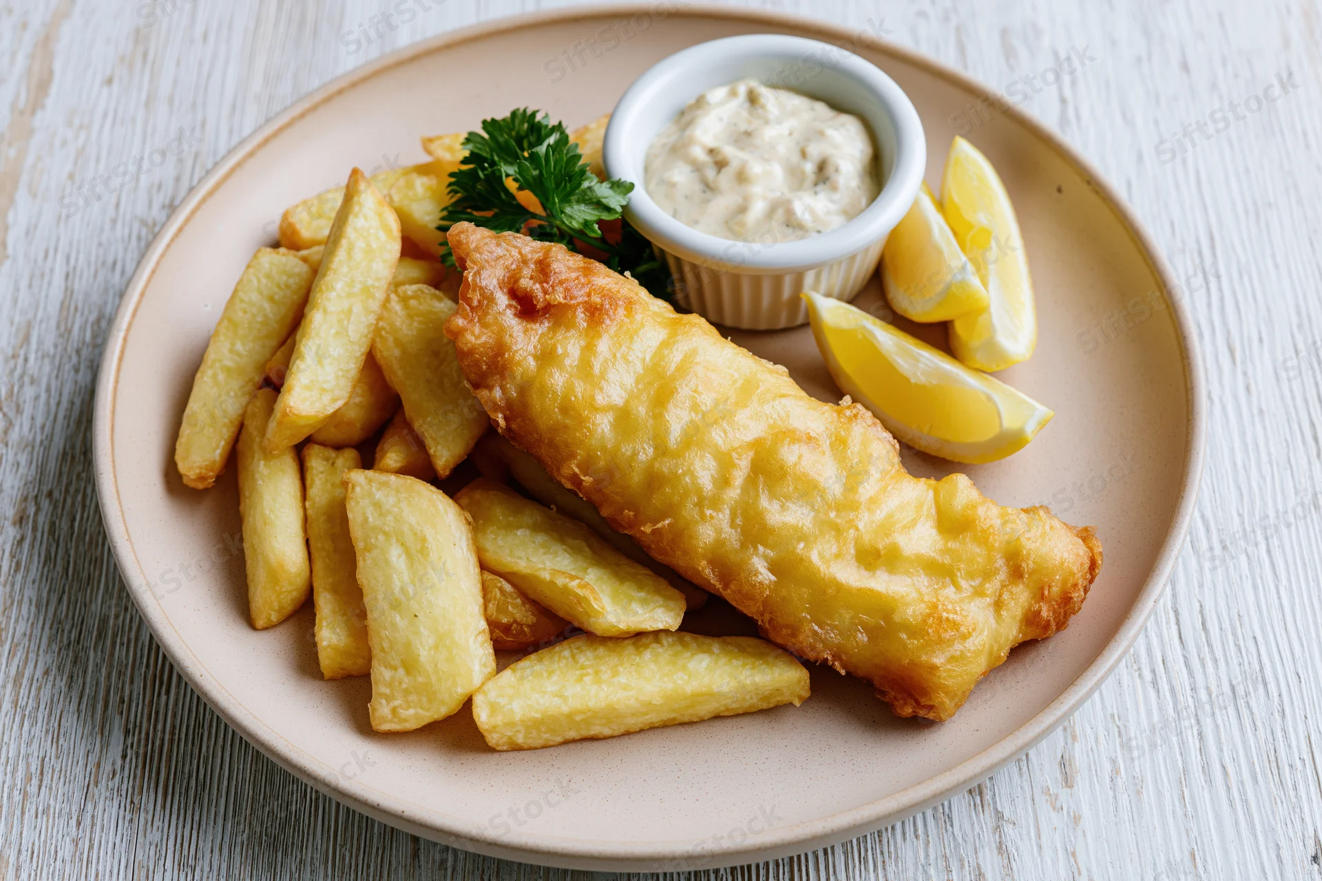 Golden fried fish and thick-cut chips on a beige ceramic plate with tartar sauce and lemon wedges