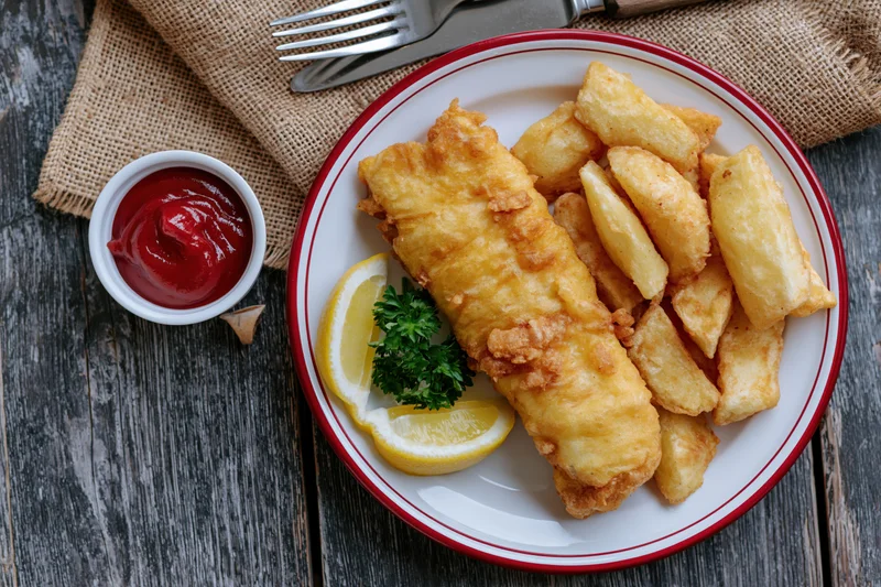 Fried fish and chips on a white plate with red rim served with lemon and parsley on a dark wooden table