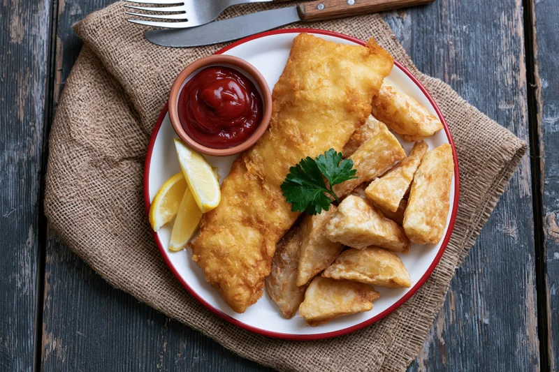 Fish and chips served on a white plate with red rim on a burlap napkin over wooden table
