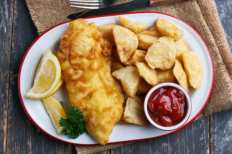 Fried fish and chips served on a white plate with red rim, garnished with lemon and parsley, on a burlap mat over dark wood table