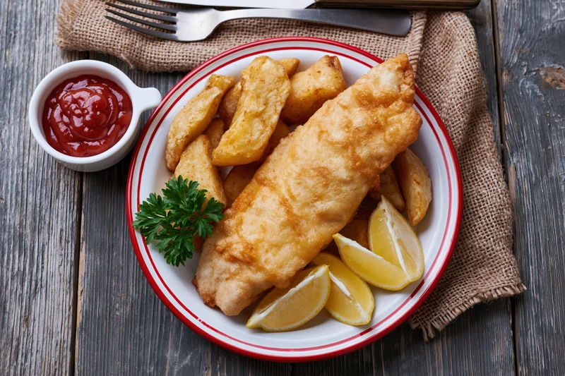 Classic fish and chips served on a white plate with red rim, garnished with lemon wedges and parsley, accompanied by ketchup, on a dark wooden table with cutlery and burlap napkin