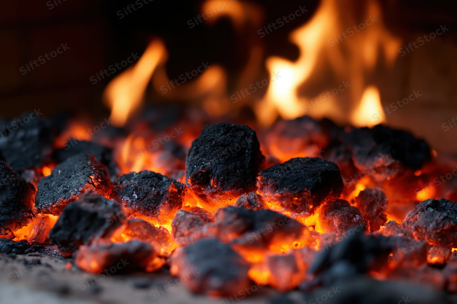 Glowing coal embers burning inside a fireplace with flames rising in the background