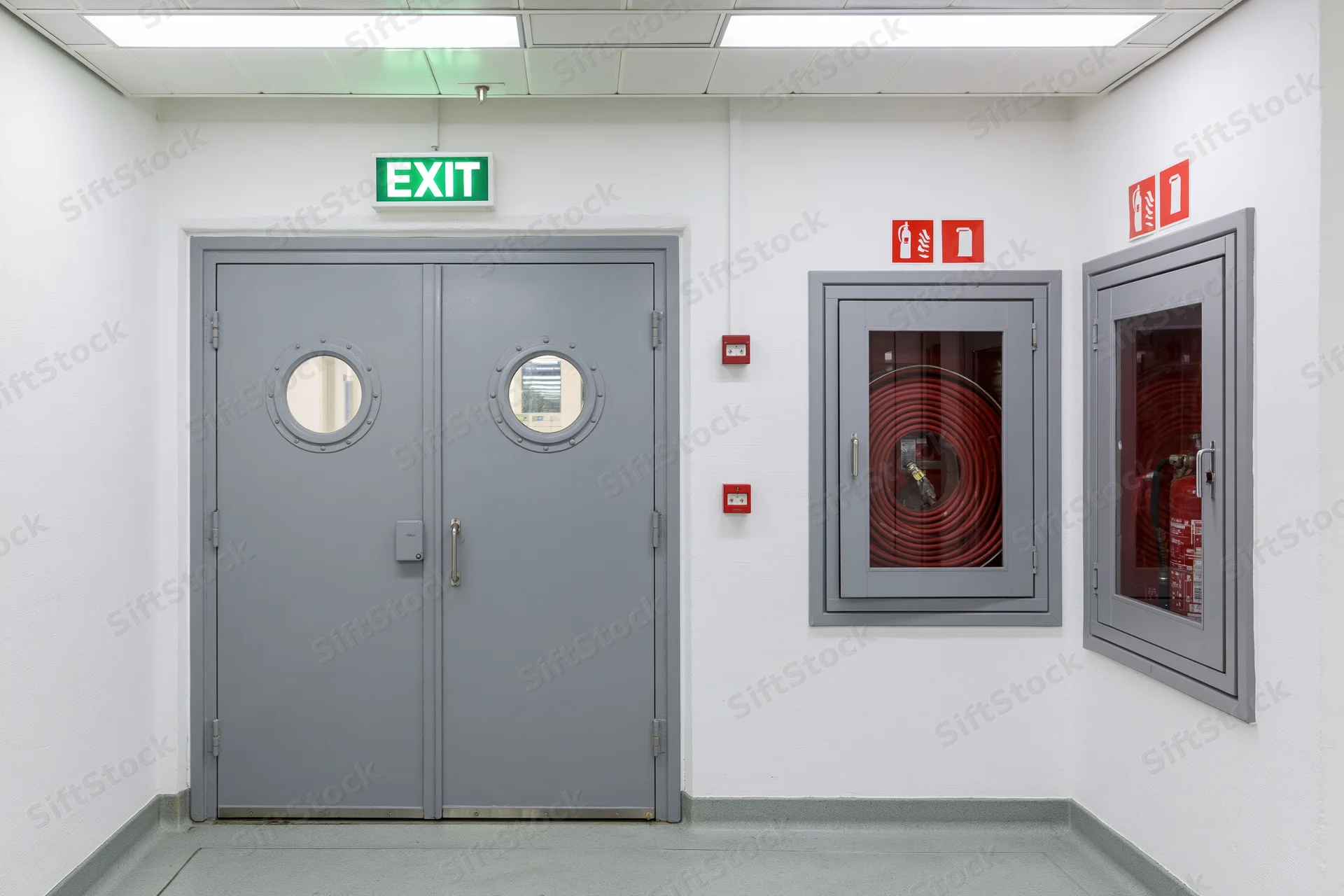 Double gray metal doors with circular windows and green exit sign in a commercial building corridor, flanked by fire extinguisher cabinets and red safety signs on white walls