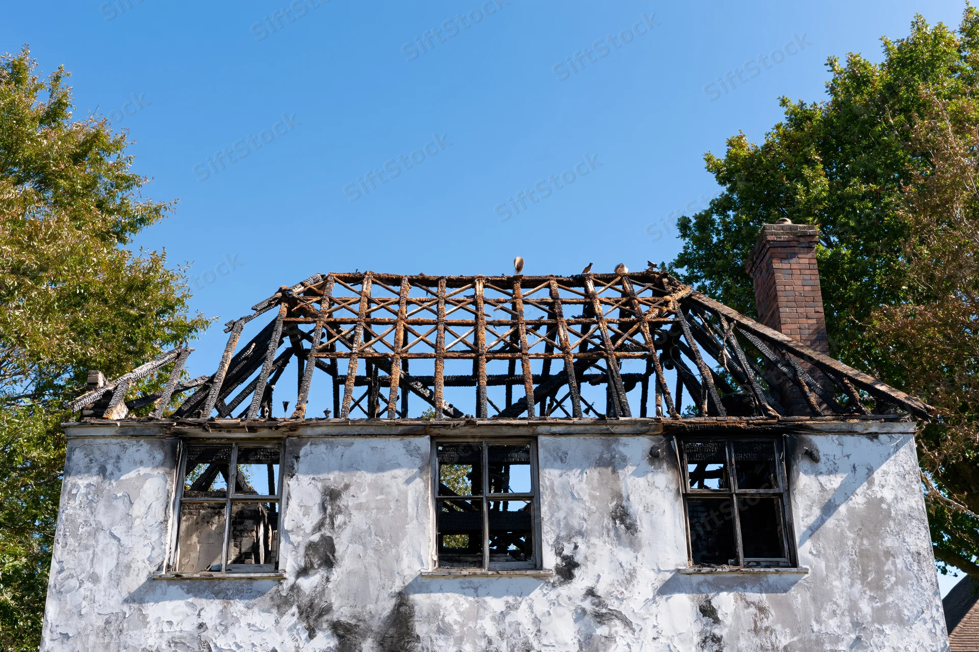Burned two-story house with collapsed roof and broken windows under clear blue sky