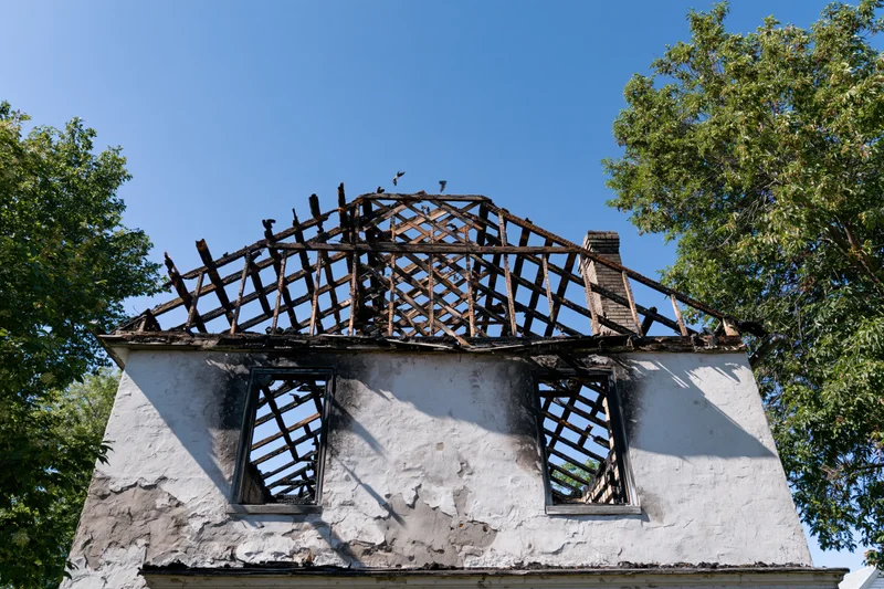 Fire-damaged house with collapsed roof structure and charred beams under blue sky