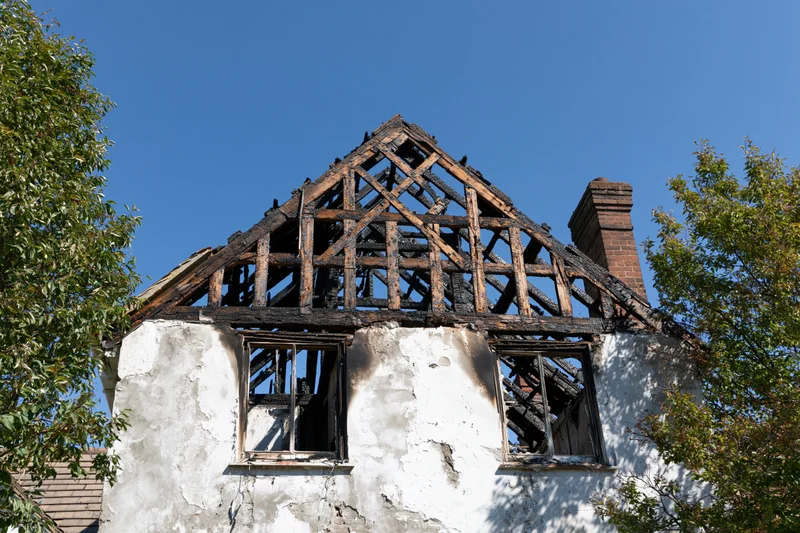 Burned two-story house with collapsed roof structure and broken windows under blue sky