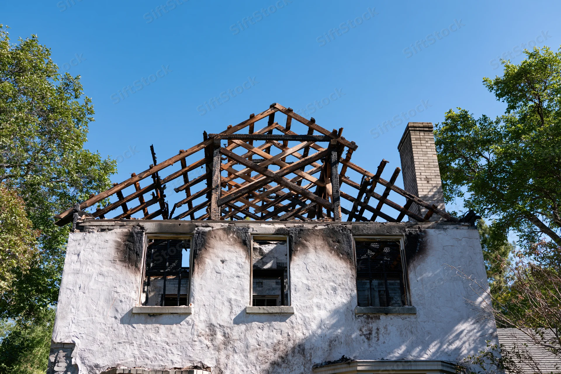 Burned two-story house with collapsed roof structure and charred exterior under clear blue sky