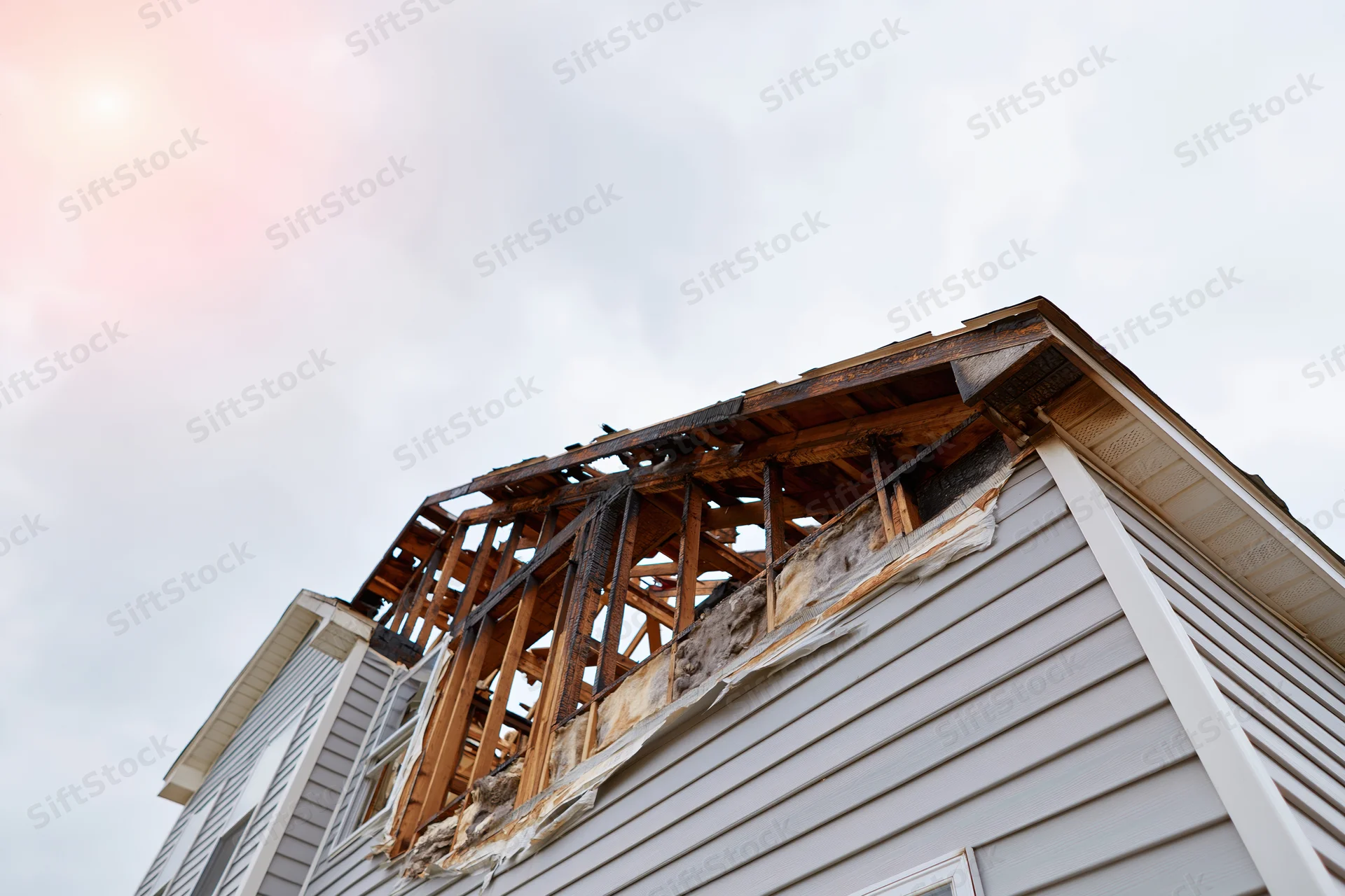 Fire-damaged two-story house with collapsed roof and exposed wooden frame under overcast sky