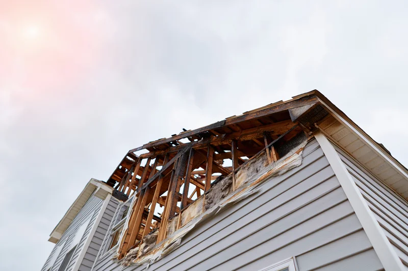 Fire-damaged two-story house with collapsed roof and exposed wooden frame under overcast sky