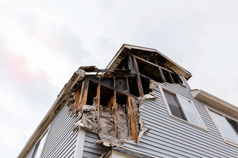 Fire-damaged two-story house with collapsed upper section and exposed framing under overcast sky