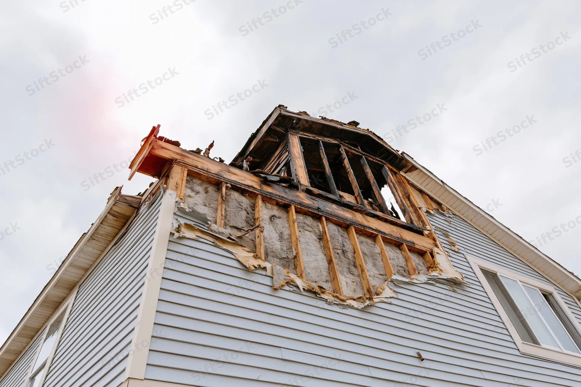 Fire-damaged two-story house with collapsed roof and exposed wooden frame under overcast sky