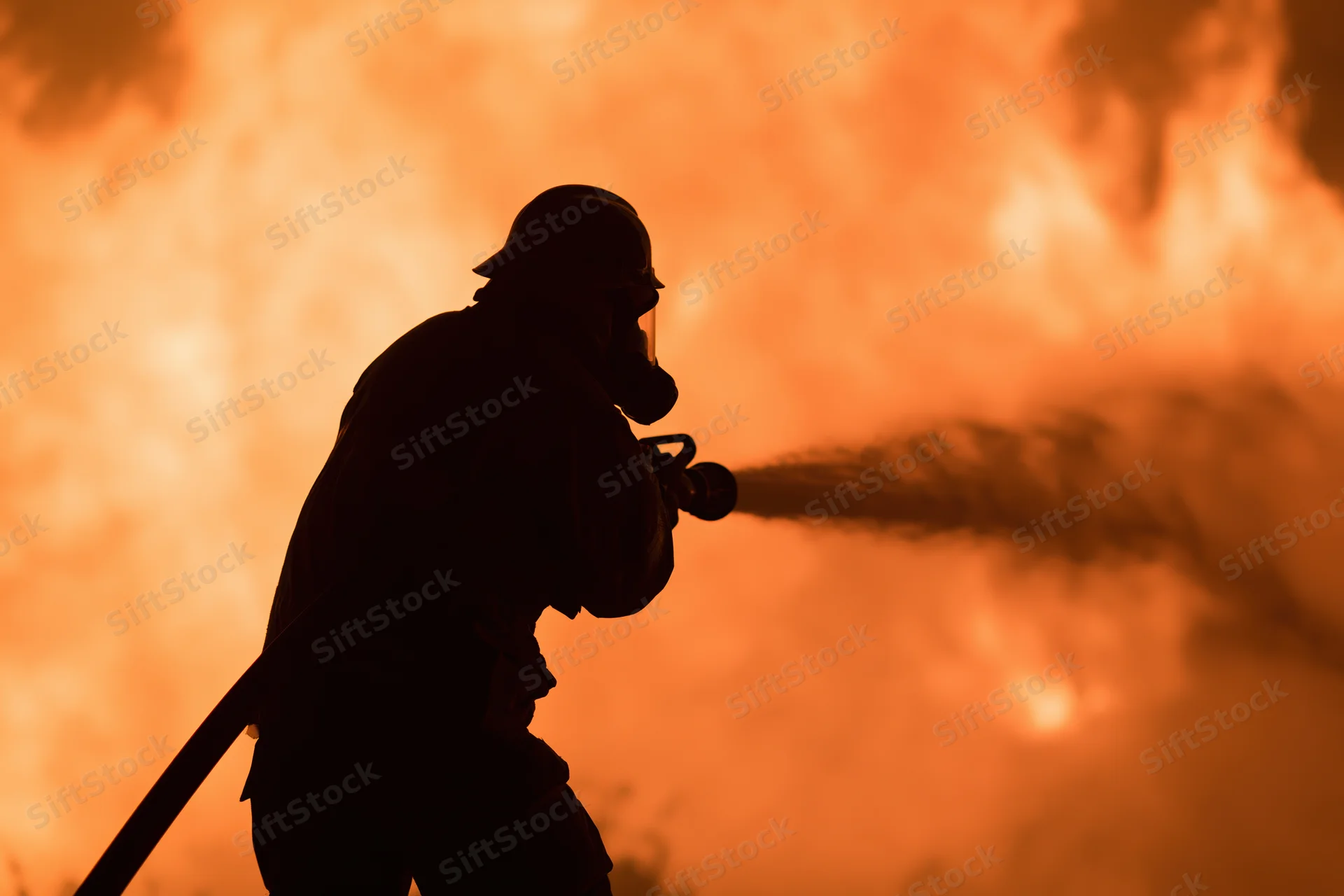 Firefighter silhouetted against a massive wall of orange flame spraying water from a hose