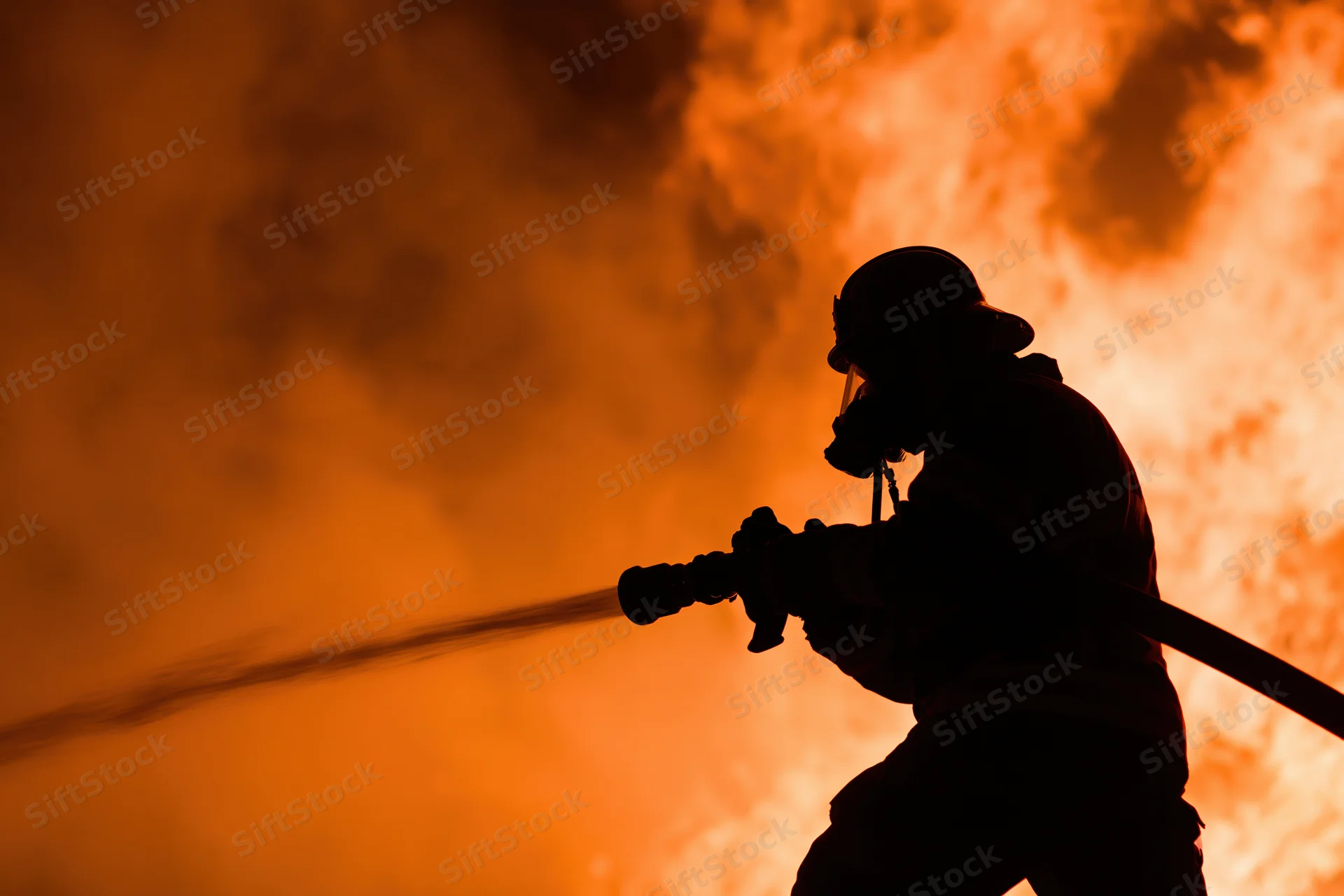 Firefighter silhouetted holding firehose against orange flames and smoke